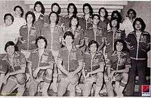 A black and white photograph of men standing in three rows, mostly wearing jackets and shorts used for basketball. Two men on the left edge of the second and back rows, and a man at the right edge of the back row are wearing white collared shirts.