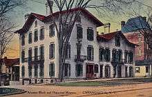 A hand-tinted postcard showing a Victorian-looking white building with many doors and windows and a pointed red roof