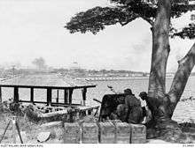 Men crouch behind the shield of an artillery gun beneath a tree overlooking a body of water