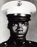 Head of a young black man staring intently at the camera, wearing a dark military jacket and a white peaked cap with a black visor.