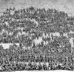 A large group of men in military uniform sit on the side of a large stone edifice.