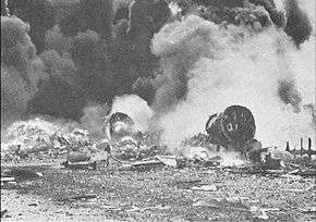 Photo of burning aircraft emitting large smoke plumes and surrounded by rubble.