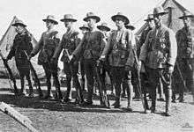 Men in military uniforms with rifles and bandoliers stand at ease in ranks