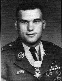 Head and shoulders of a white man with short dark hair wearing a military jacket with a star-shaped medal hanging loosely from a ribbon around his neck.