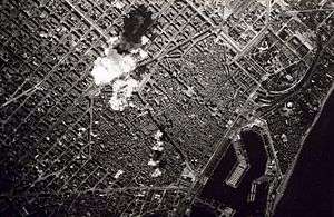 Black-and-white photo of the city from high above. Smoke from a bomb can be seen