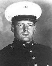 Head and shoulders of a young white man wearing a white peaked cap with a black visor and a dark military jacket with bright buttons down the center.