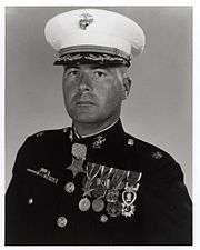 Head and shoulders of a middle-aged white man wearing a white peaked cap with oak leaf decorations on the visor and a dark military jacket with two rows of medals hanging from ribbons pinned to his chest and another medal hanging from his neck.