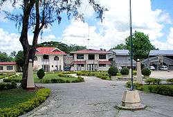 Five broad paved paths meet at centre.  Between the paths are grassed areas with low hedge borders.  The area at left holds a large tree.  No people to be seen. In the background, perhaps 50 metres away, are several two-storey white-faced buildings.  They could be residential, they could be administrative.  In the foreground is a flagpole, no flag.