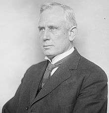 A white-haired man, sitting, wearing dark jacket, patterned tie, and white shirt