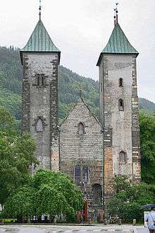 Stone  Romanesque church with two  towers and a  lower, pitched roof entrance hall between them. The towers have slightly curved  bronze roofs.