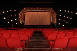 interior shot of red velvet seats facing a gilded decorated proscenium arch flanked by decorative wall lights