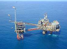 An oil platform constructed in two main parts sits in a calm blue sea. The larger section at right has a helideck, the smaller at left is flaring gas from two stacks.