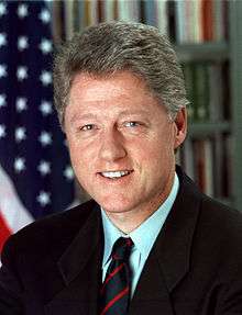 A presidential photograph of a Caucasian man wearing a suit with a black tie. An American flag and bookshelf are in the background.