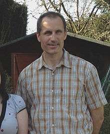 Man with oval head and crew-cut dark hair stands outdoors wearing large-checked brown and white shirt