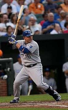 A man in a grey baseball uniform with a blue helmet holds a baseball bat while preparing for a pitcher to throw the ball to him.