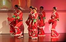 Six women in red gowns with white trim on a wooden floor against a white background performing a dance