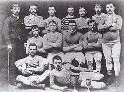 A group of thirteen men, twelve in association football attire typical of the late nineteenth century, and one in a suit holding a cane. Two trophies are in front of the players.