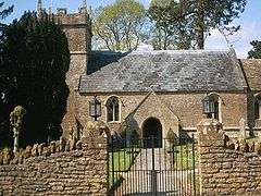 Small stone building with square tower partially obscured by trees. In the foreground is a stone wall with metal gates.