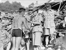 A large crowd of men in uniforms with slouch hats. A woman in uniform with skirt and flat bush hat presents a small trophy to a man in a swim suit.