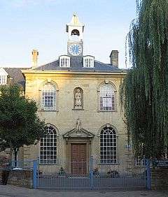 Yellow stone building with central 2-storey block with arched windows, hipped roof and clock tower