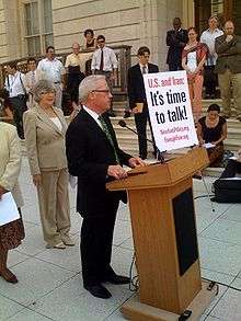 Barr stands outside at a podium with the words "U.S. and Iran: It's time to talk" written on a poster standing upright in the front, surrounded by listeners