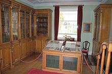 Photograph of a laboratory, with glass-encased, wooden bookcases on two walls and a window on the third. There is a display case in the middle of the room.