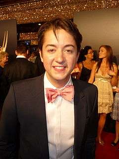 A man with dark hair, wearing a black suit, including a pink bow tie and white shirt.