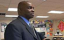 A man, wearing a black jacket with blue shirt, is standing in the middle of a classroom.