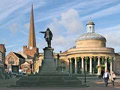 Statue of figure with outsretched arm. To the left a tall church spire and to the right a circular building with columns.