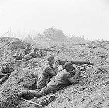 A black-and-white photograph of a battle scene in which two Vickers machine guns are firing from behind cover of a pile of rubble. Each machine gun is crewed by two men, a firer and an observer with a pair of binoculars. Another soldier is looking on from behind the crews