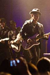 A man fully dressed in black with a necklace playing guitar for a crowd with a bass player and another guitar player in the back.