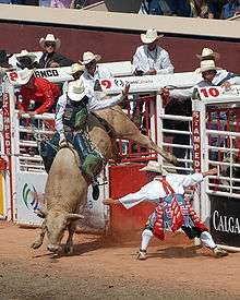 A man attempts to hang onto a rope tied around a bucking bull, while a rodeo clown and several cowboys look on.