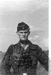 A man wearing a camouflage military uniform, side cap and a pair of binoculars around his neck. His cap has an emblem in shape of a human skull and crossed bones.