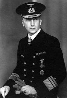 A man wearing a peaked cap and dark military uniform with an Iron Cross displayed at the front of his uniform collar. He is holding gloves in his hands.