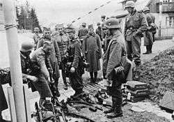 A black and white photograph of German soldiers watching other soldiers laying down their rifles in a pile.