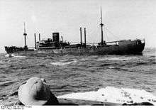 A dark-coloured merchant ship at sea. The head of a person wearing a sun helmet is visible in the foreground