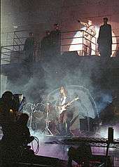 A concert stage in front of a wall with 2 levels. Five men stand on a balcony, including Roger Waters, who is saluting with his arm and is lit by a spotlight. On the lower level is a drum kit and a man playing guitar.