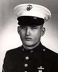 Head and shoulders of a young white man wearing a white peaked cap with black visor and a dark military jacket with bright buttons down the center of the chest.