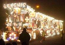 Night time photograph of lorry and trailer illuminated with thousands of lamps to make pictures. In the foreground and to the side are pedestrians.