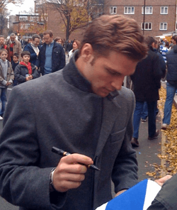 A man wearing a dark grey woollen coat signs an autograph outside for a fan.