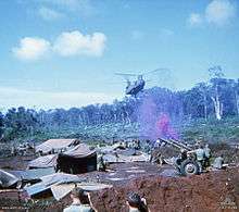 Colour image of a large two-rotored helicopter flying past a line of trees and preparing to land in an open area cleared of vegetation. In the foreground are a large number of tents, hootchies and ground sheets, while a number of soldiers are standing around an artillery piece. Purple smoke is rising from the position.