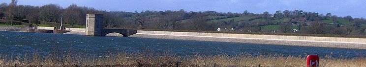 Photograph showing the white coloured dam running horizontally across the picture with water and vegetation in the foreground and hills behind