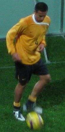 A man with dark hair in a yellow, long-sleeved top kicking a football.