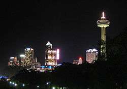 A spike tower with a pod is lit along its length on the right. Other buildings show interior lights in this night scene. The foreground is a deep black sky.