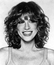 A black and white photo of a blond, curly haired woman wearing a tank top is seen smiling.