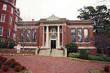 A one-story brick building with grey concrete stairs in the center leading to a door with a column on either side of it. There are three long windows on each side of the building.