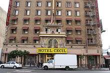 The lower five stories of a tan brick building in a city. Its lower windows have awnings; there is a fire escape on the right. At the street there is a large shelter over the entrance with "Hotel Cecil" on it in black type on a yellow background. A small truck and car, both white, are parked on the street in front.