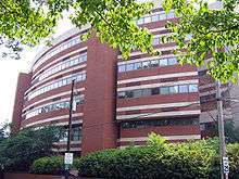 A round, six-story brick building with rows of windows. At its base, there are green bushes and a power line. In the foreground above, there are branches of a tree with green leaves.