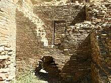 Inside daytime view of a ruined and ceiling-less rectangular room. Tawny-beige stacked sandstone bricks compose walls rising from brush-covered ground. The several walls visible in the image are up to perhaps a dozen feet in height. In the wall immediately at center, a triangle-shaped entrance several feet high leads to an adjacent chamber behind. The upper part of the same wall, shaped like an inverted-triangle, has fallen away or otherwise been removed, revealing a rectangular doorway leading to yet another concealed room. At left and right are two similar walls perpendicular to the one at middle.