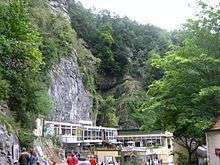 Exposed limestone cliffs with some tress and vegetation above white buildings with large windows.